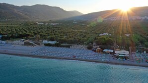 A coastal area with a beach, rows of sun loungers, and a mountainous backdrop.