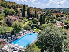 A pool with lounge chairs and umbrellas, surrounded by greenery and stone buildings.