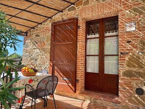 A stone building with a wooden door and window, a table with a fruit basket, and a chair.