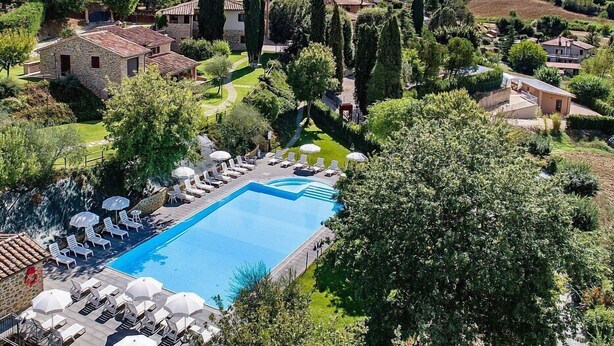 A pool with lounge chairs and umbrellas, surrounded by greenery and stone buildings.