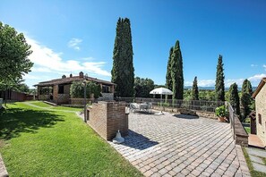 A patio area with a brick floor, outdoor furniture, and tall cypress trees.