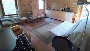 A cozy room with a stone wall, wooden ceiling, a bed, a table with chairs, a small kitchen area, and a radiator.
