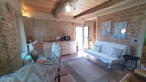 A rustic room with a stone wall, wooden ceiling beams, a sofa, a dining table, and a kitchen area.