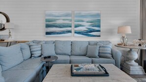 A living room with a light blue sofa, two paintings of the ocean, a coffee table with a tray, and a lamp on a side table. - Private Beach | 1 minute walk to Beach! | Beautifully updated 1 bedroom condo | Sleeps 6 | Wi-Fi (Santa Rosa Beach)