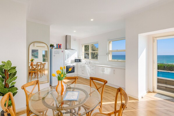 A modern kitchen with white cabinets, a dining area with a glass table, and a view of the sea through large windows. - Glenelg Beach House With Private Beachfront Pool (Glenelg North)
