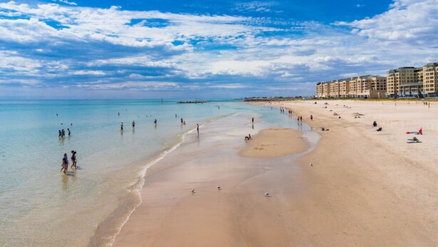 A wide beach with clear blue water, people walking along the shore, and a row of buildings in the background. - Glenelg Beach House With Private Beachfront Pool (Glenelg North)