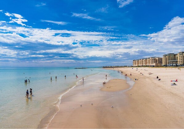 A wide beach with clear blue water, people walking along the shore, and a row of buildings in the background. - Glenelg Beach House With Private Beachfront Pool (Glenelg North)