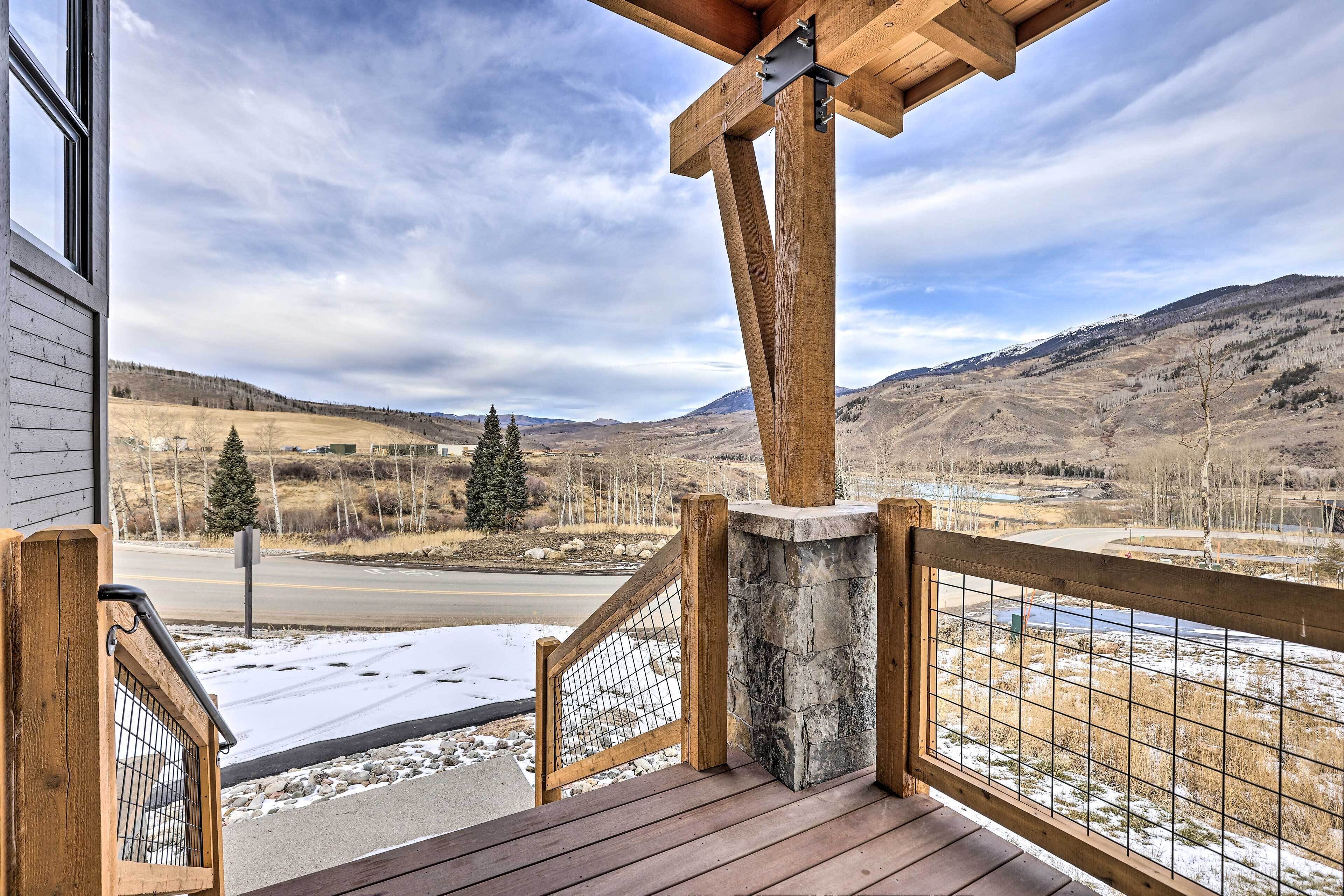 A wooden deck with a stone pillar, a metal railing, and a view of a snowy landscape with mountains and a road.