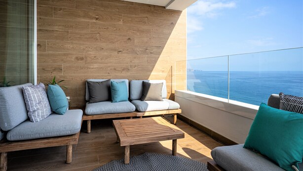 A modern balcony with a wooden floor, grey sofas with patterned cushions, and a wooden coffee table.