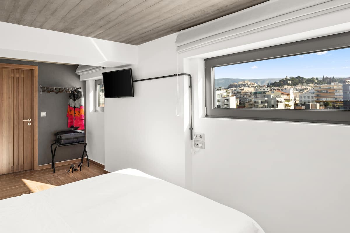 A modern hotel room with a large bed, a television mounted on the wall, a window with a city view, and a wooden door.