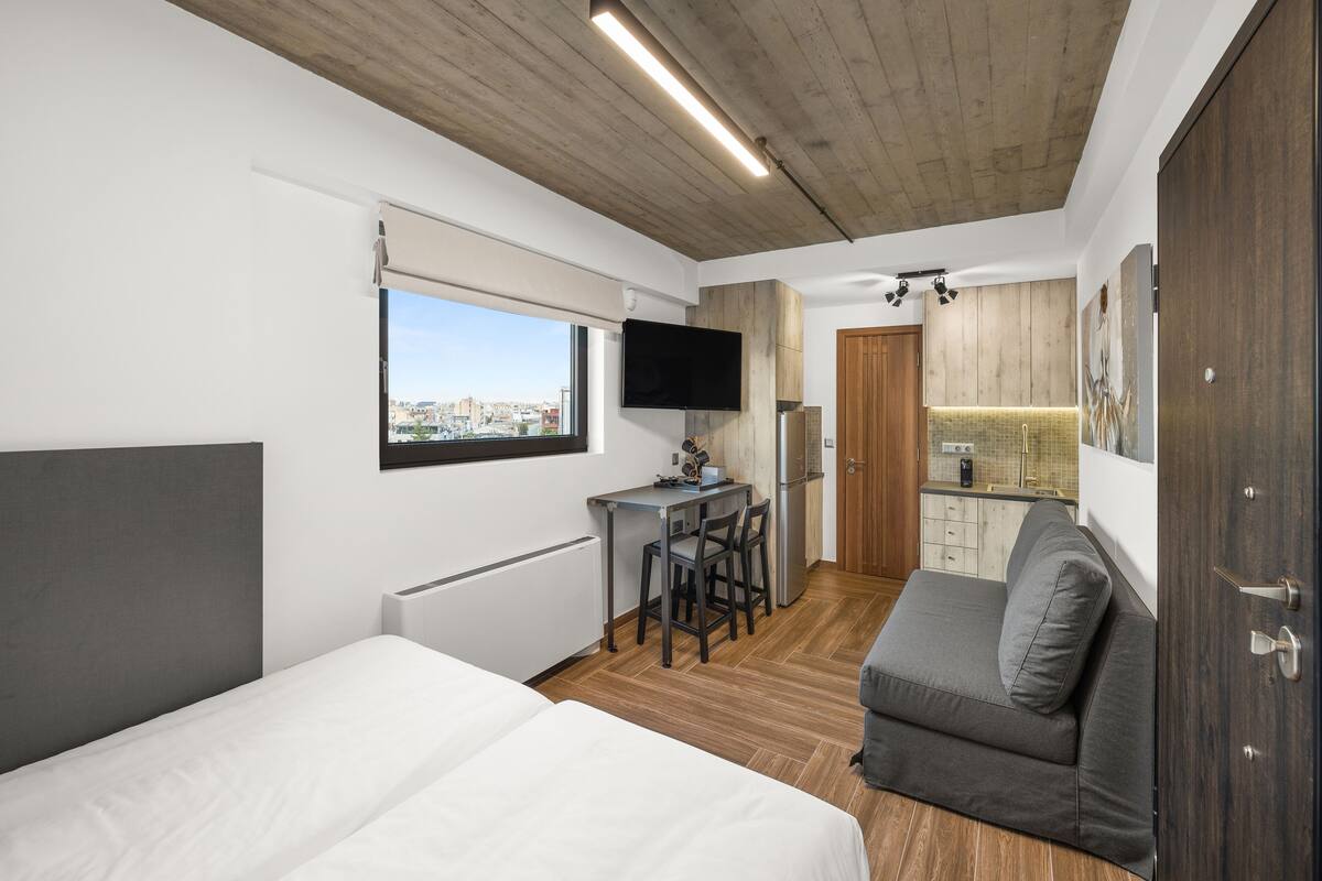 A compact room with a bed, a small kitchenette, a seating area, and a TV mounted on the wall.