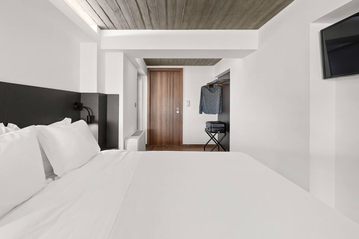 A modern hotel room with a large bed, a wooden door, a TV mounted on the wall, and a clothing rack.