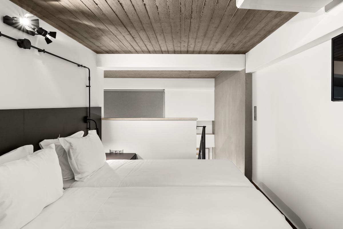 A modern hotel room with a wooden ceiling, a large bed with white linens, a bedside table, and a wall-mounted light fixture.
