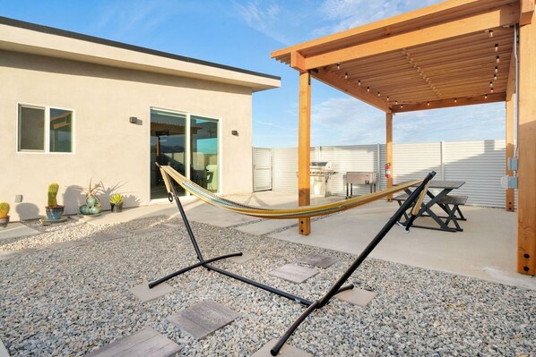 A hammock under a wooden pergola in a backyard with a patio table and chairs, surrounded by potted plants.