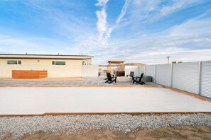 A covered outdoor area with a fire pit and seating, adjacent to a building with a garage door. - Sun Oro - Hot Tub, BBQ, Fire Pit and Gameroom (Joshua Tree)