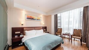 Une chambre d’hôtel avec un lit, des tables de chevet, un bureau, des chaises, un tableau et de grandes fenêtres.