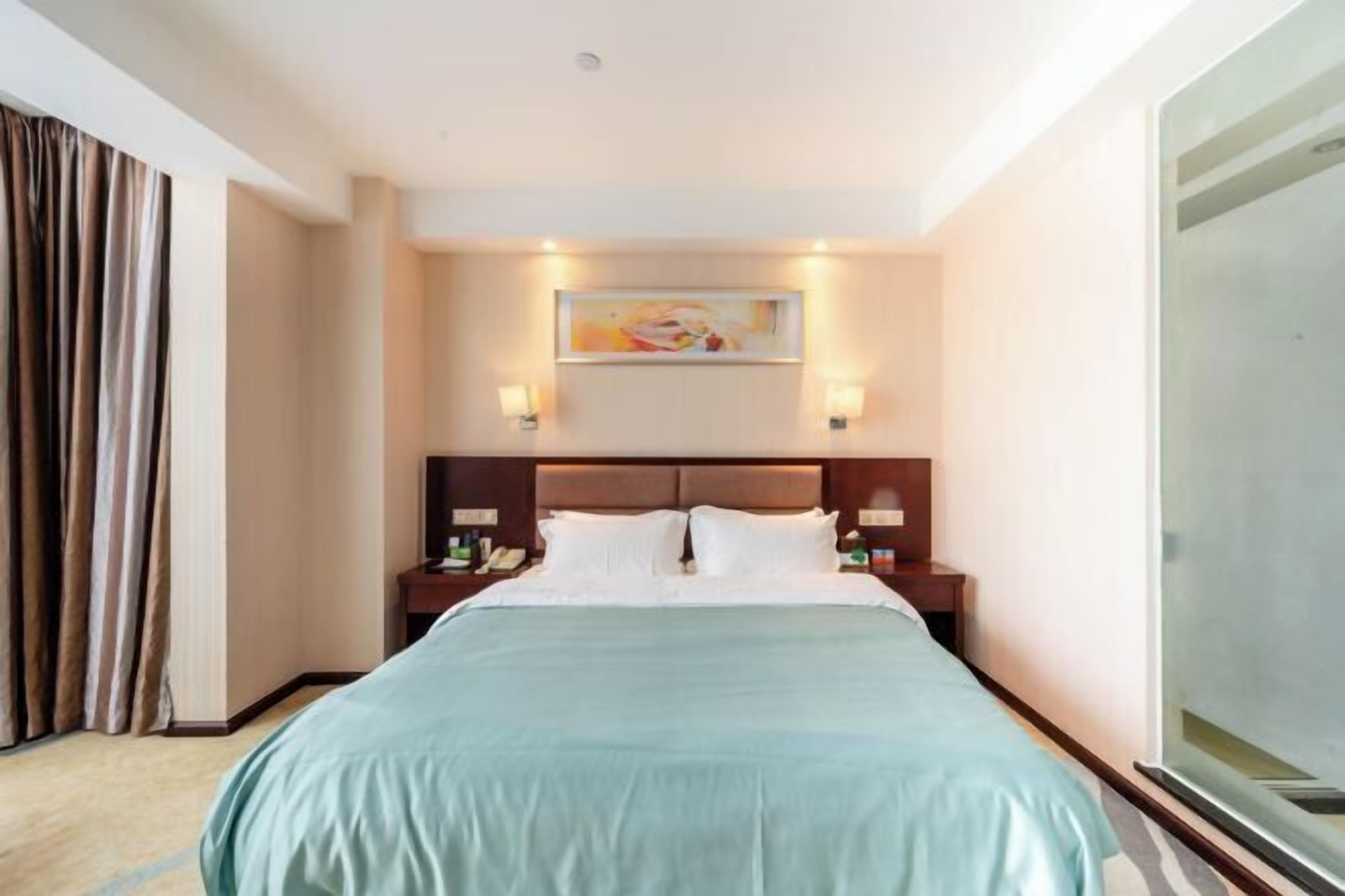 A hotel room with a large bed, two bedside tables, a painting on the wall, and a window with curtains.