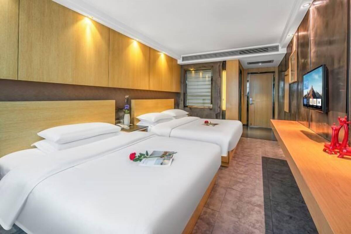 A hotel room with two beds, a wooden headboard, a TV on the wall, and a reception desk with a red sculpture.