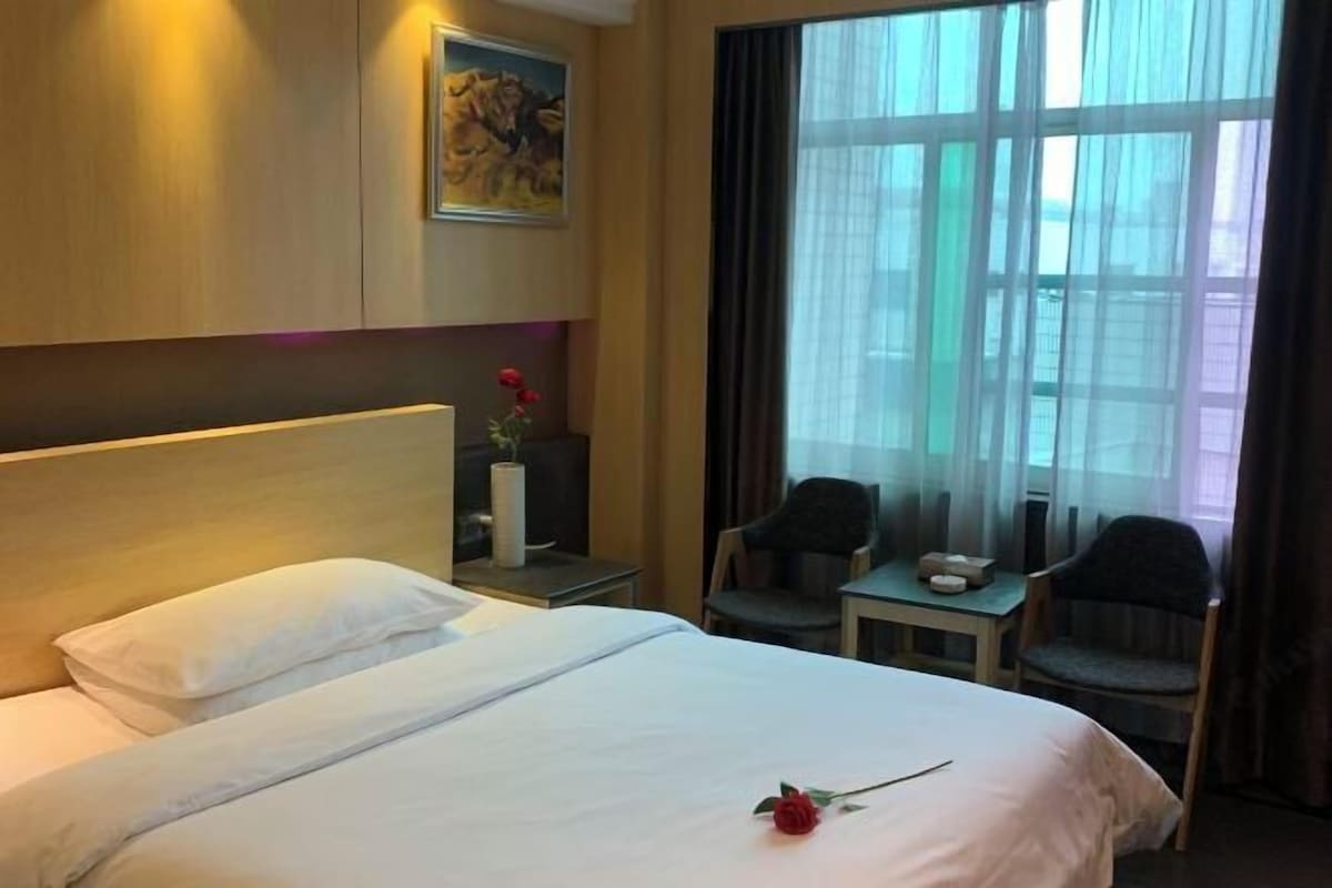 A hotel room with a bed, a small table with chairs, a painting on the wall, and a window with curtains.