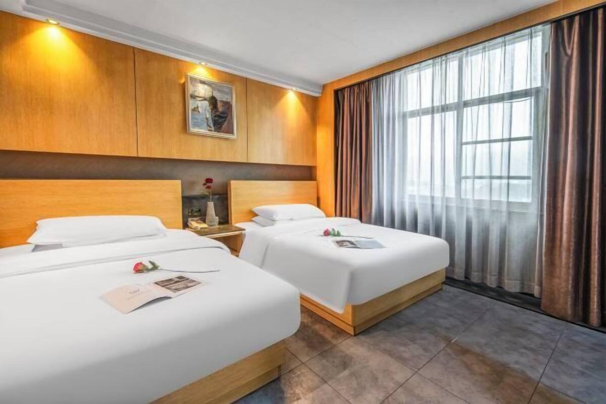 A hotel room with two beds, wooden headboards, a large window with curtains, and a painting on the wall.