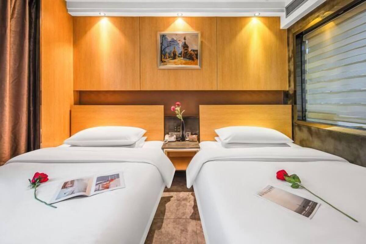 A hotel room with two beds, a wooden headboard, a painting, a nightstand, and a window with blinds.