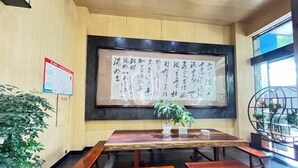 Una mesa de madera con bancos, una gran obra de caligrafía en la pared y una planta en maceta.