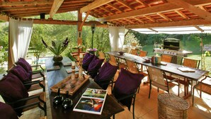 An outdoor patio with wooden tables and chairs, a grill, and a view of the surrounding greenery.