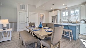 A modern kitchen with a central island, white cabinetry, and a dining area with a wooden table and cushioned chairs. - Kure Beach Townhome: Short Walk to the Beach! (Kure Beach)