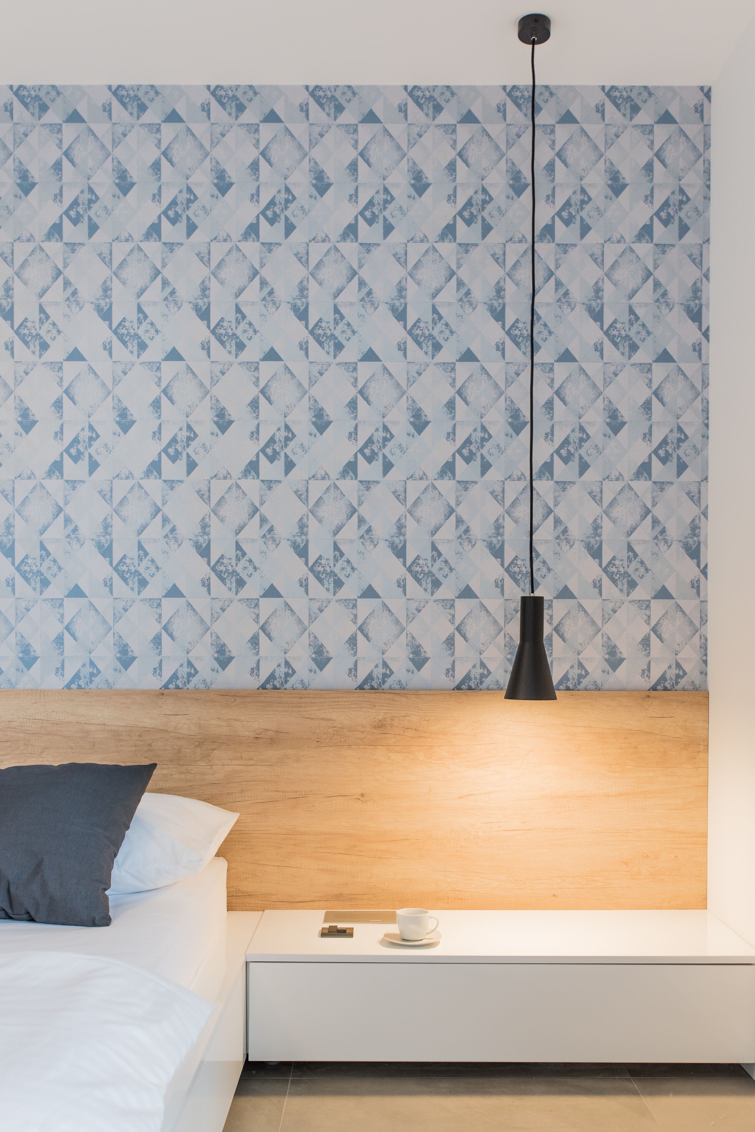 Une chambre avec un papier peint à motifs, une tête de lit en bois, une table de chevet avec une tasse et une lampe suspendue.