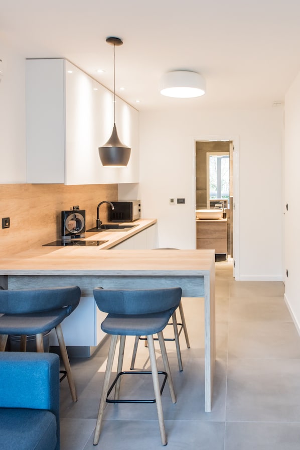 A modern kitchen with a wooden countertop, a built-in microwave, and a pendant light. There are two blue bar stools and a wooden table.