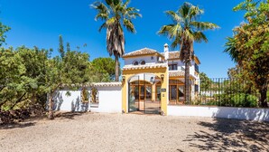 Un bâtiment à deux étages avec une façade jaune, une entrée fermée par un portail et des palmiers en arrière-plan.