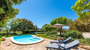 Une piscine entourée de verdure et dotée d’un sol en pierre, avec des chaises longues et un parasol.