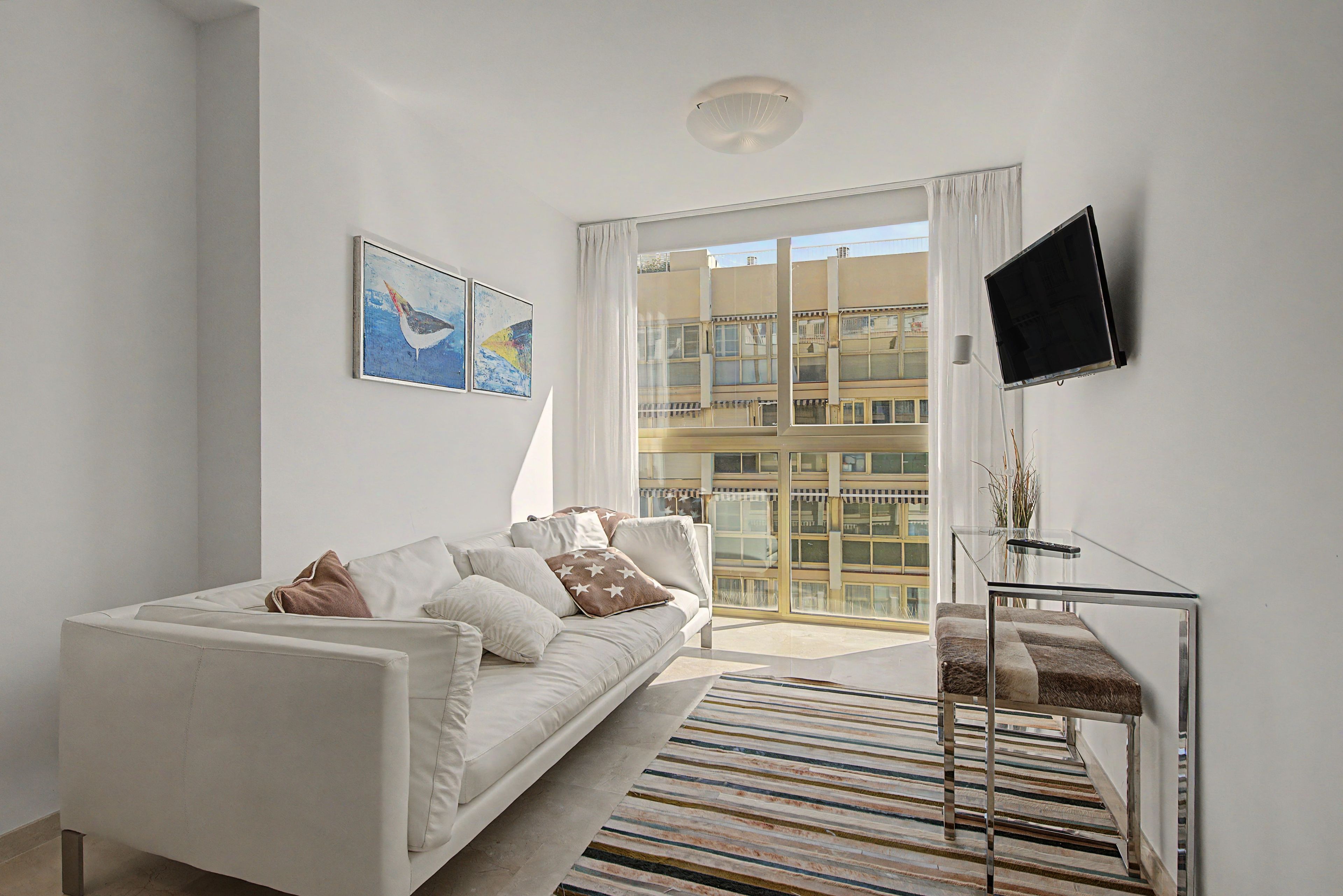 A modern living room with a white sofa, a flat-screen TV mounted on the wall, and a large window with a view of a building.