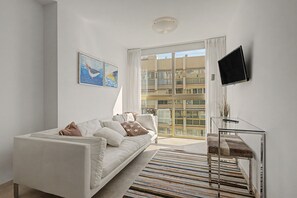 Un saló modern amb un sofà blanc, una televisió de pantalla plana penjada a la paret i una finestra gran amb vista a un edifici.