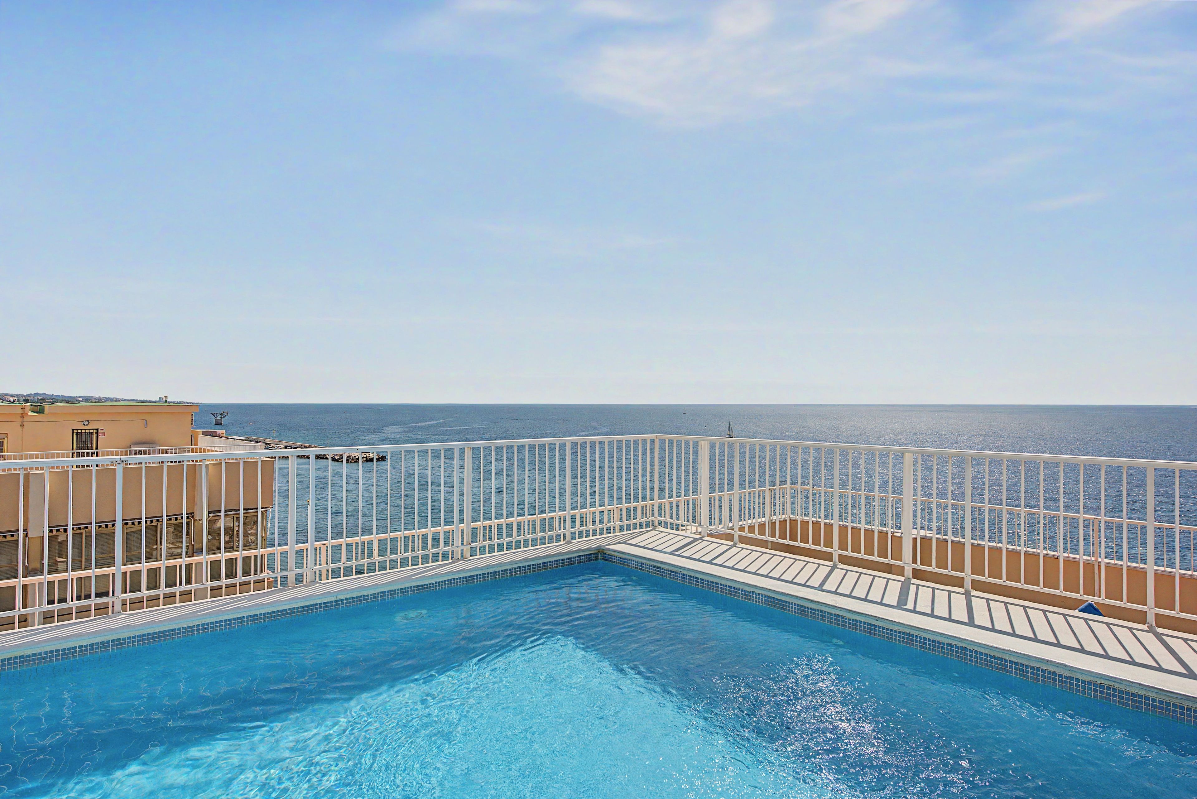 A pool with a white railing, surrounded by a building with a view of the sea.
