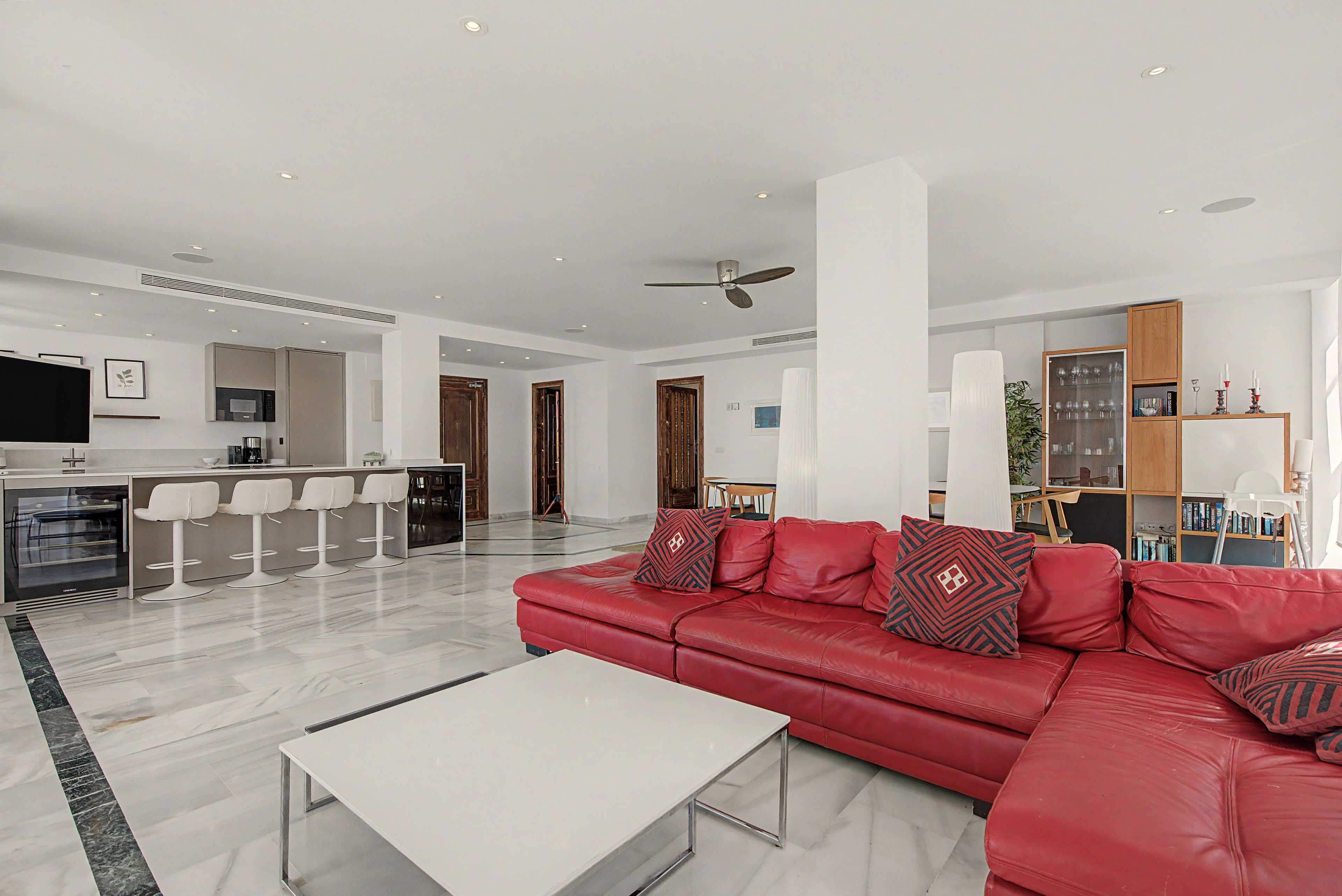 A modern living room with a red leather sofa, a white coffee table, and a kitchen area with a microwave and bar stools.