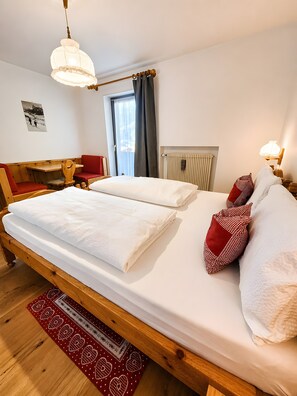 Un lit double avec du linge de lit blanc, une tête de lit en bois, un tapis à motifs rouges et blancs, un bureau en bois avec une chaise, et une fenêtre avec des rideaux.