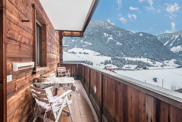 Un balcon en bois avec une table et des chaises, donnant sur un paysage montagneux enneigé.