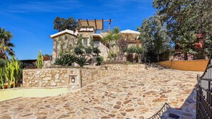 A stone-paved courtyard with a stone wall, a wooden pergola, and a building with a balcony.
