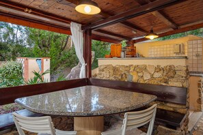 Un espace extérieur abrité, aménagé pour les repas, avec un mur en pierre, un plafond en bois et une table entourée de chaises.