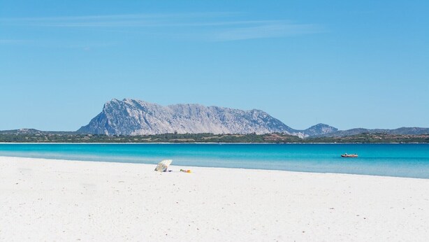 A beach with white sand, a clear blue sea, and a mountain in the background.