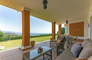 Un espacio al aire libre techado con mobiliario de mimbre, una mesa con fruta y vistas al paisaje.