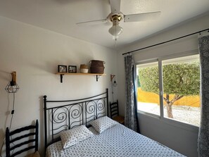 A bedroom with a bed, a ceiling fan, a window with curtains, a shelf with decorative items, and a wall-mounted lamp.