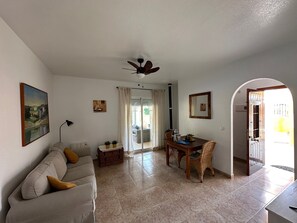A living room with a sofa, a dining table with chairs, a ceiling fan, and a view of the outside.
