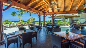 An outdoor patio with wooden tables and chairs, overlooking a beach with palm trees and the ocean. - 🏖️Garden View Waikoloa Condo w/ A/C + Ocean Club Access (Waikoloa)