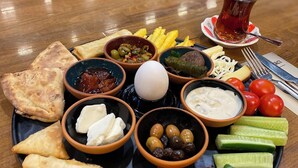 Uma mesa com diversos alimentos, incluindo pão, azeitonas, queijo e um copo de chá.