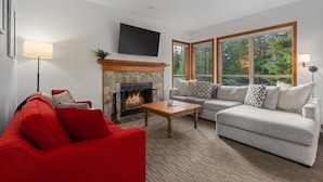 A modern living room with a stone fireplace, a red sofa, a grey sofa, and large windows offering a view of the forest. - 2BR | Painted Cliff | Ski in/out | Communal Hot Tub | Wood Burning Fireplace | Free Parking (Whistler)