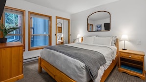 A bedroom with a bed, bedside tables, a dresser, a mirror, and a window with blinds. - 2BR | Painted Cliff | Ski in/out | Communal Hot Tub | Wood Burning Fireplace | Free Parking (Whistler)