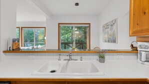 A kitchen with a large window, a built-in sink, wooden cabinets, and a coffee maker. - 2BR | Painted Cliff | Ski in/out | Communal Hot Tub | Wood Burning Fireplace | Free Parking (Whistler)