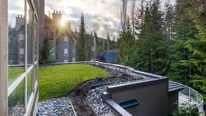 A modern building with a glass facade, a landscaped garden with a pebble path, and a view of a forested area. - 2BR | Painted Cliff | Ski in/out | Communal Hot Tub | Wood Burning Fireplace | Free Parking (Whistler)